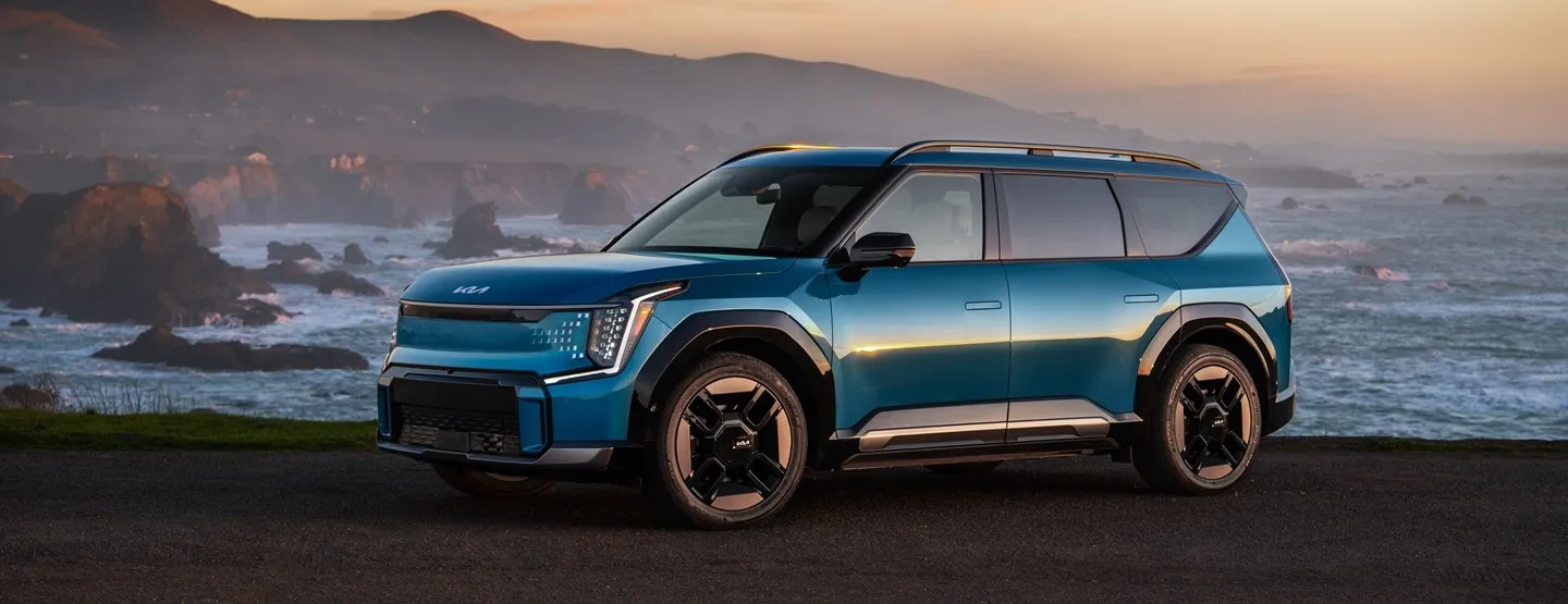 Blue Kia electric SUV parked on a coastal road with rocky cliffs and ocean waves in the background during sunset.