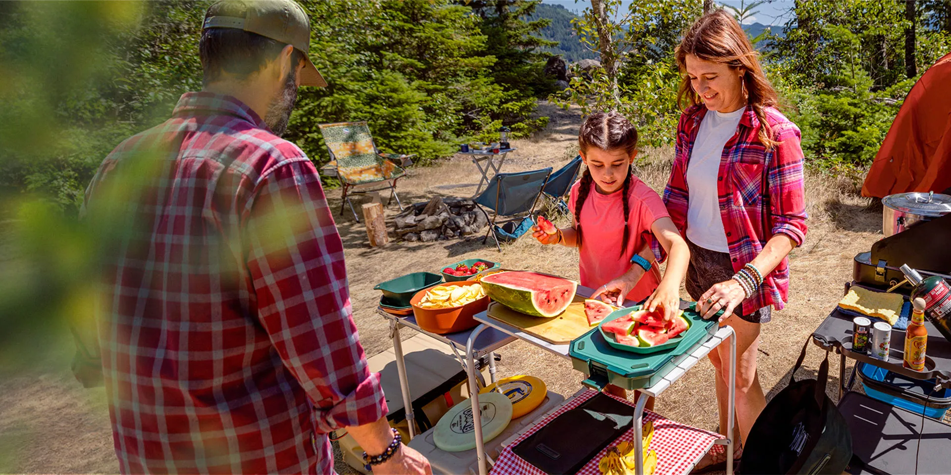 Family at a campsite preparing and serving sliced watermelon at a portable campsite table surrounded by camping gear and chairs.