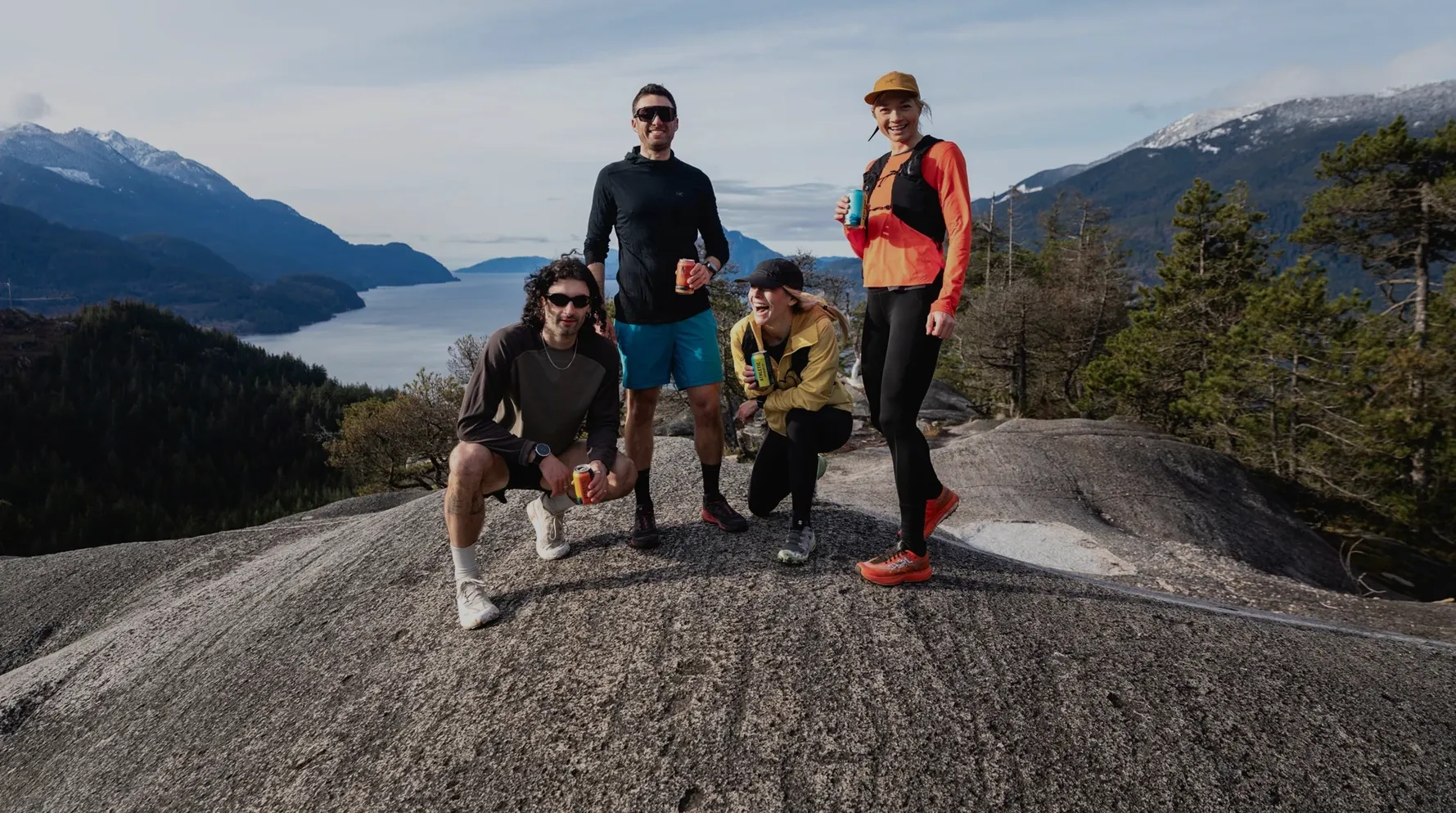 Group of four hikers holding drinks on a rocky overlook with mountains, forest, and a lake in the background.