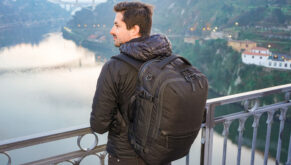 A man wearing a premium black travel backpack on his back is looking over a bridge