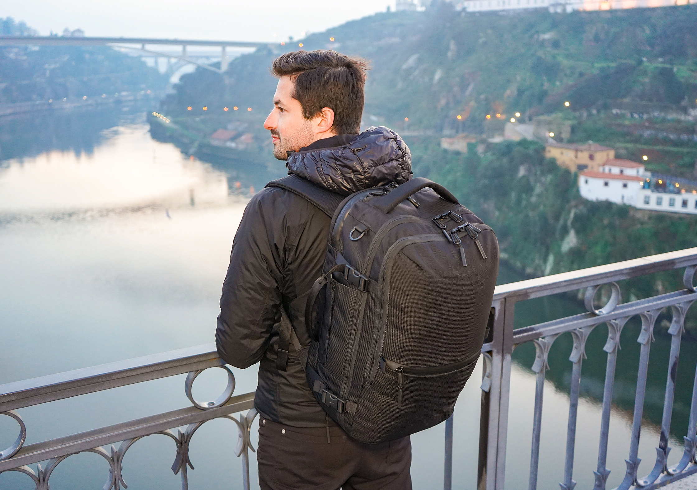A man wearing a premium black travel backpack on his back is looking over a bridge